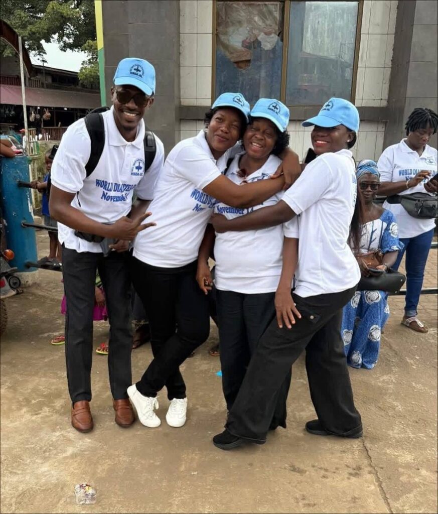 Group of people in white shirts and blue hats hugging and smiling to the camera with Help Madina in Sierra Leone for World Diabetes Day