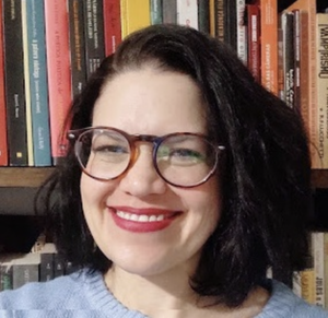 Headshot of Renata with glasses, lipstick and shoulder length dark hair smiling at the camera in front of a bookself