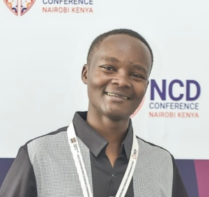 A heashot of Chasia, a black man with short hair in a black shirt and a lanyard smiling at the camera