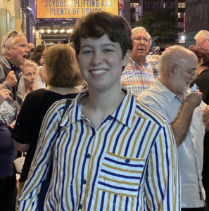 Headshot of Arden a white person in a striped shirt and shaggy brown hair smiling big at the camera