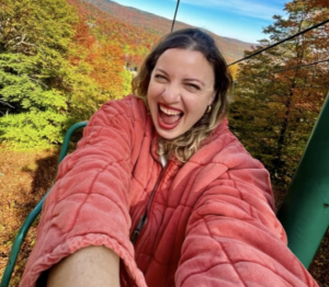 Courtney in a pink puffy on a ski lift with fall foliage behind her smiling big at the camera