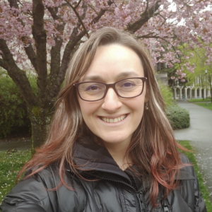 Jenny, a white person with glasses and red tips on her brown hair smiles at the camera with a tree in full bloom behind her