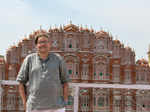Ernest in a light blue shirt stands in front of a fancy red building