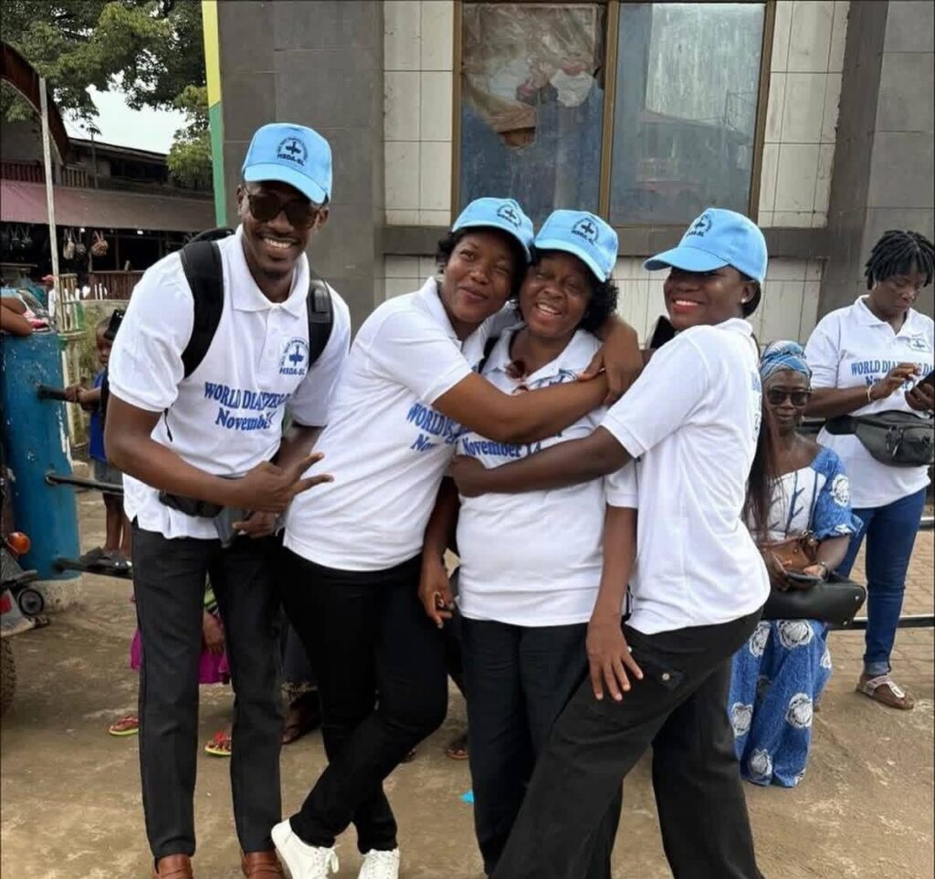 Group of people in white shirts and blue hats hugging and smiling to the camera with Help Madina in Sierra Leone for World Diabetes Day