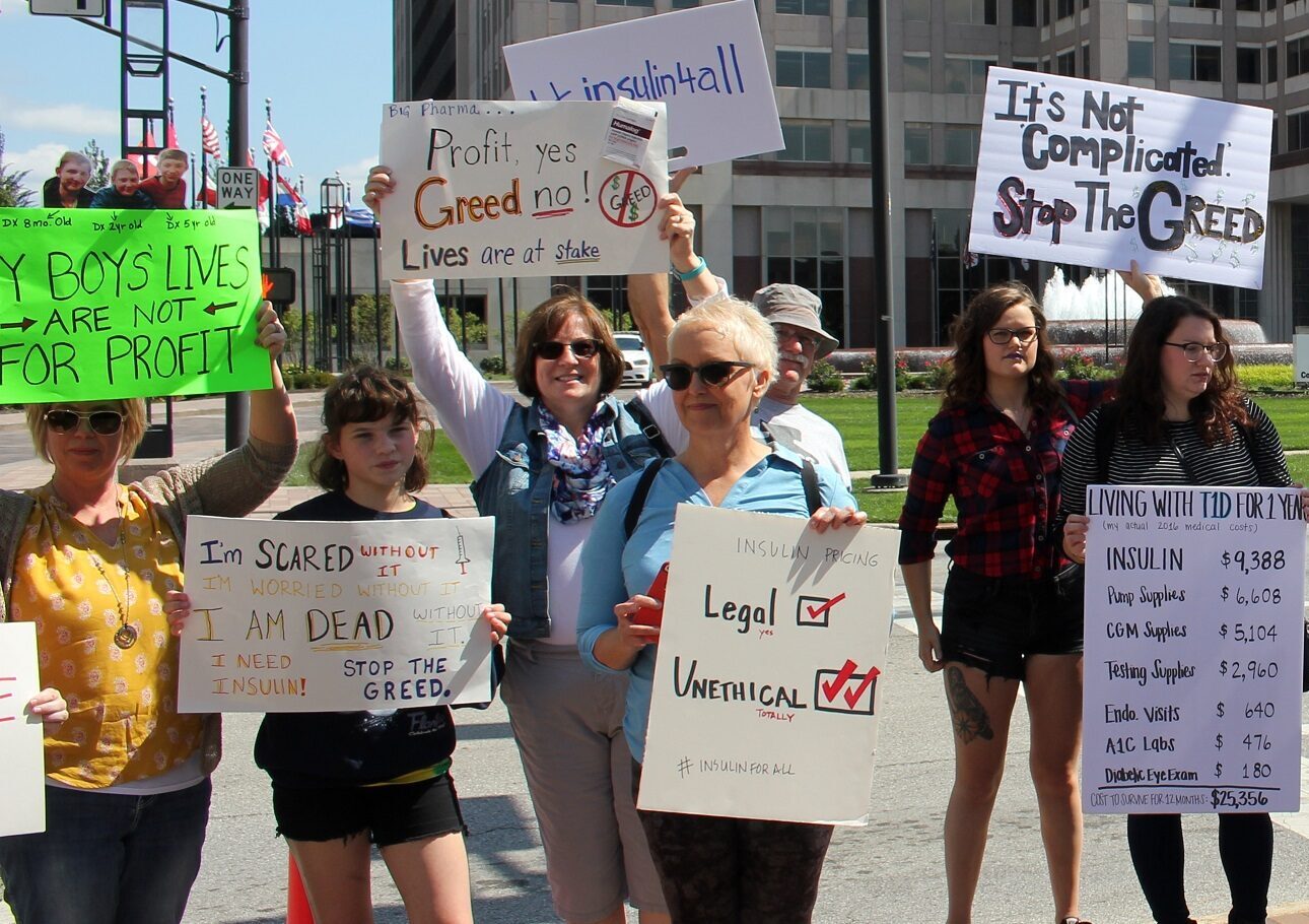 Protesters outside a pharmaceutical company headquarters holding handmade signs about insulin pricing and the annual cost of living with type 1 diabetes.