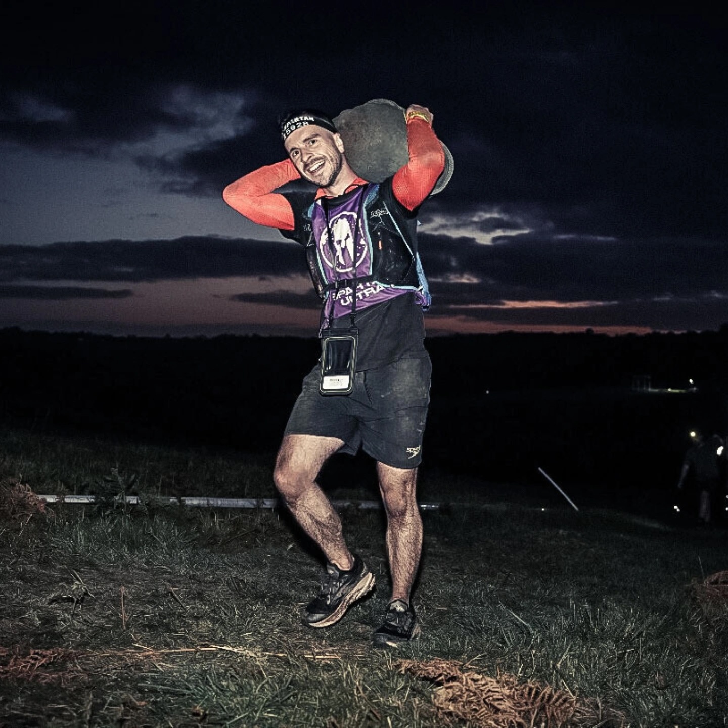 A white man in sports gear in the dark hauls a large sandbag behind his neck