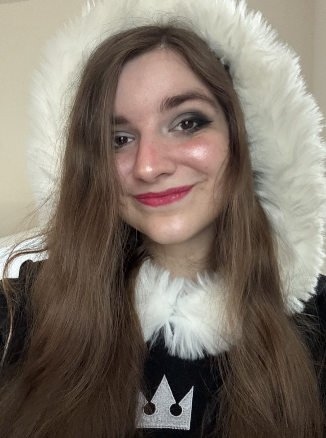 A white person with long brown hair and lipstick smiles at the camera in a white fur hood