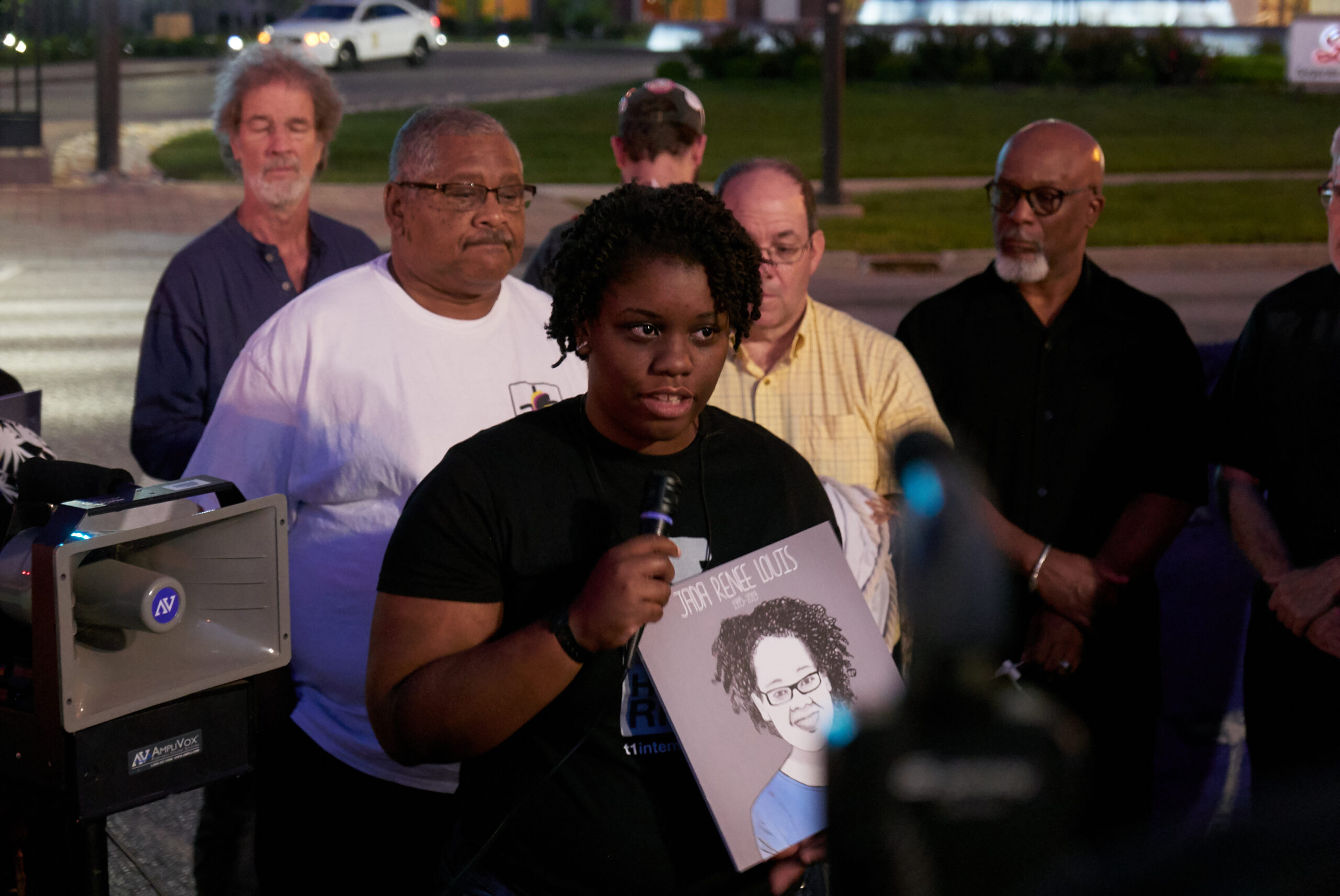 Jasmine speaking at T1International's vigil. Mic in hand, holding a drawing of her sister who died from insulin rationing