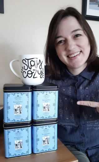 A photo of Liz smiling and pointing to a tower of 4 tins of Harney Tea with a mug balanced on top