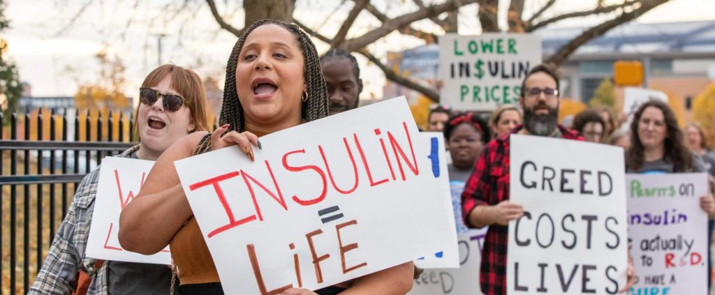 An advocate at a rally holding a hand-lettered "Insulin = life" sign, surrounded by marchers with "Insulin is a human right" placards
