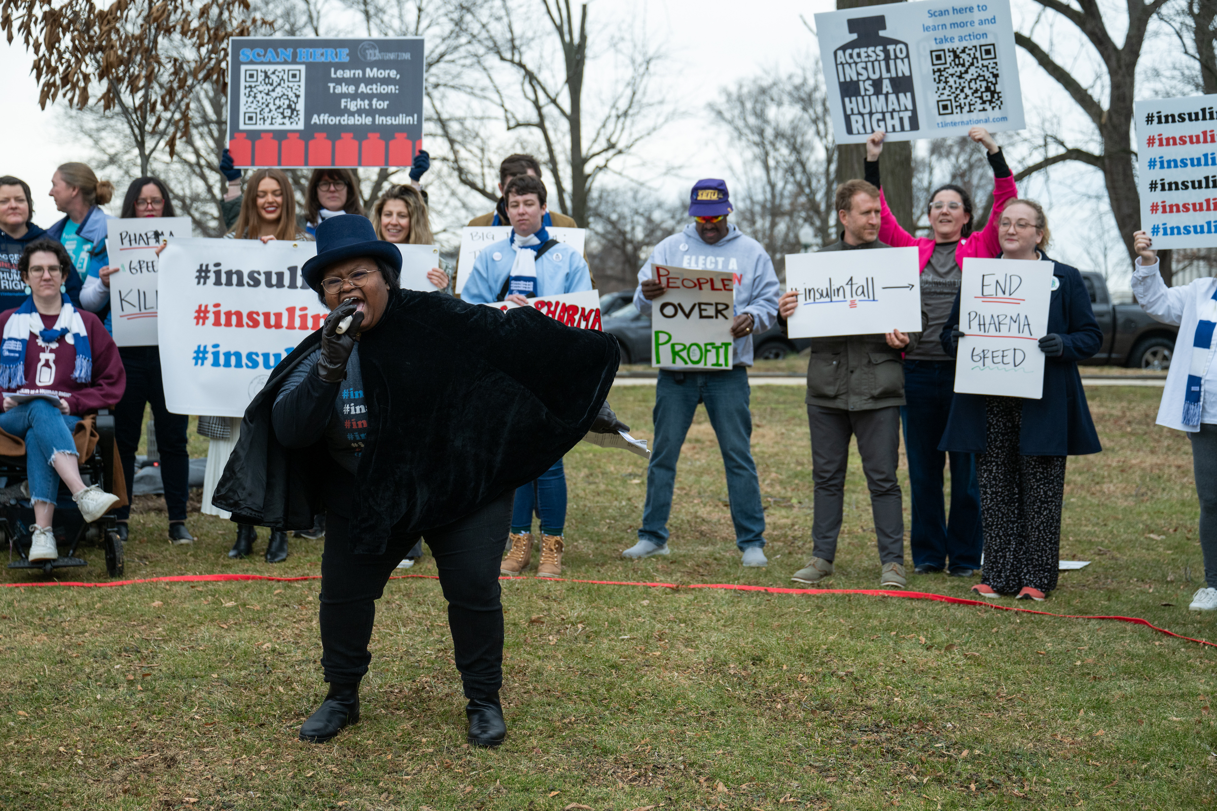 Tracy in a top hat and a cape speaking at the Circus of Pharma Greed event in front of people with signs and banners